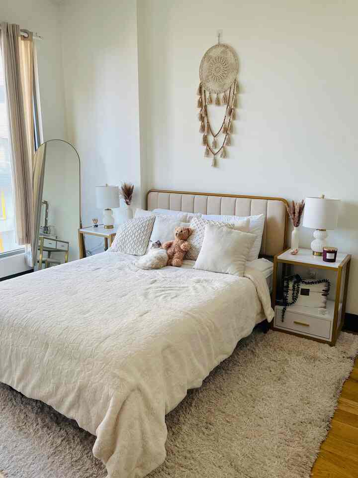 A white and beige toned bedroom featuring a central bed with nightstands and a dreamcatcher hanging on the wall, creating a simple and natural atmosphere