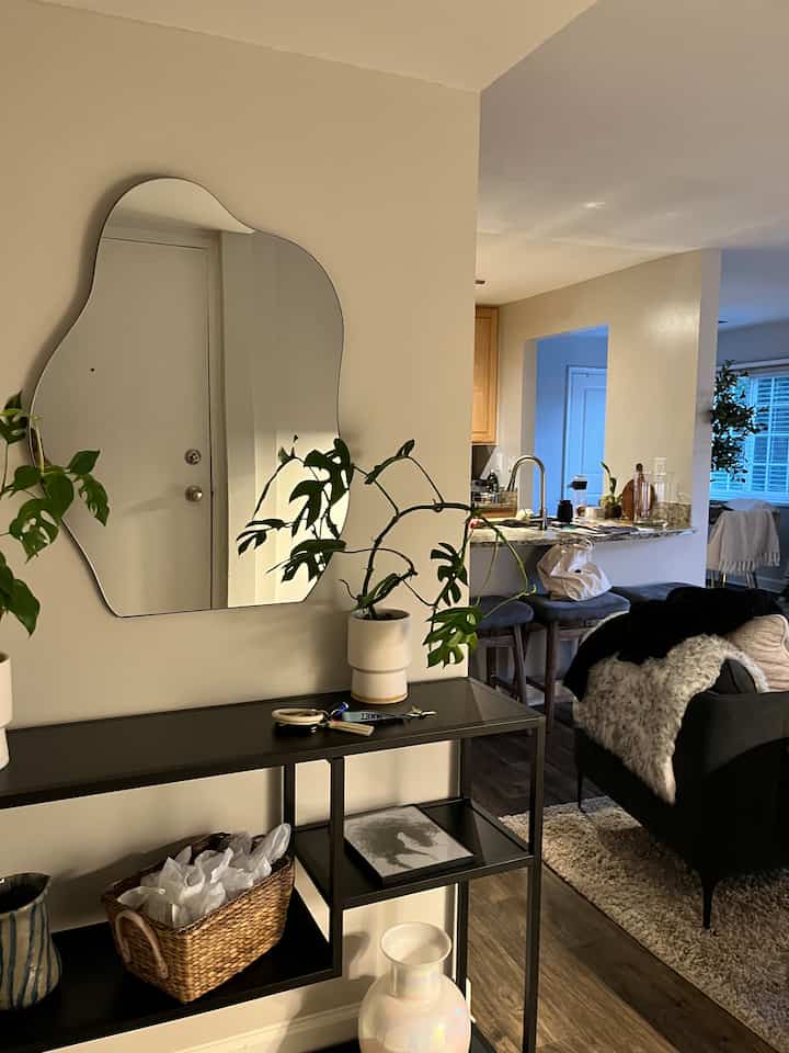 Natural color entrance featuring a wavy mirror and console table, adjacent to a living room with wood tone flooring and sofa
