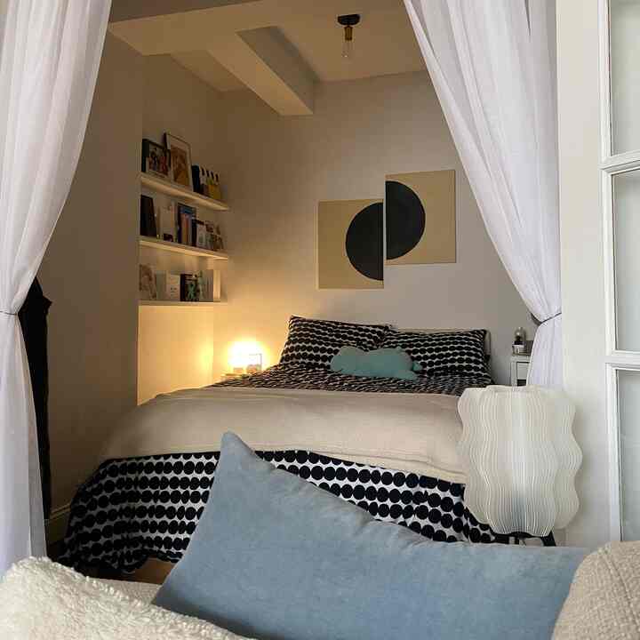Modern bedroom in white and black tones featuring a central bed with patterned bedding, wall shelves on left, and warm lighting creating a cozy atmosphere