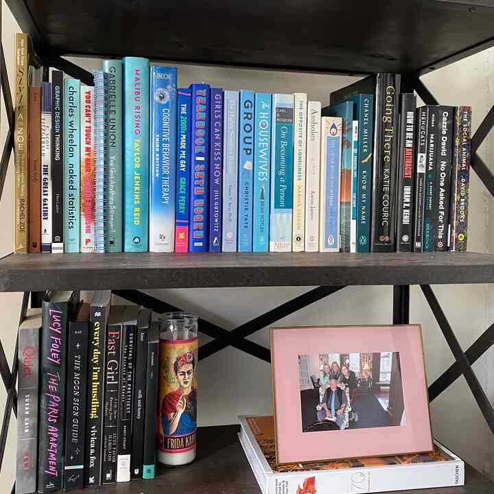 Natural brown bookshelf filled with color-organized books, featuring a Frida Kahlo candle and a pink picture frame in a cozy reading corner