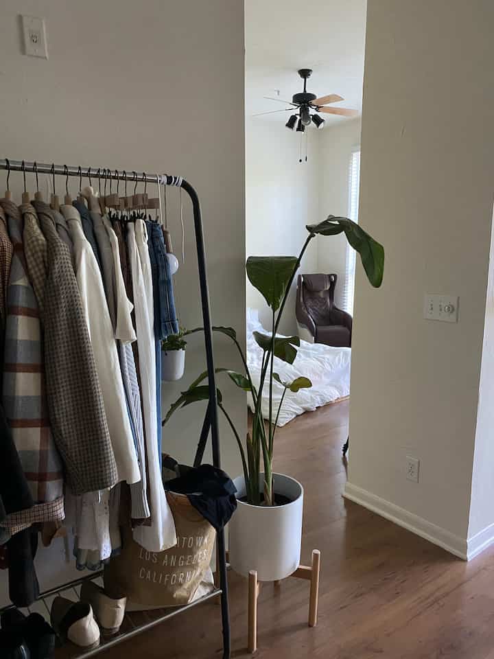 White wall and wood tone floor studio apartment featuring clothing rack and bedding with a natural atmosphere