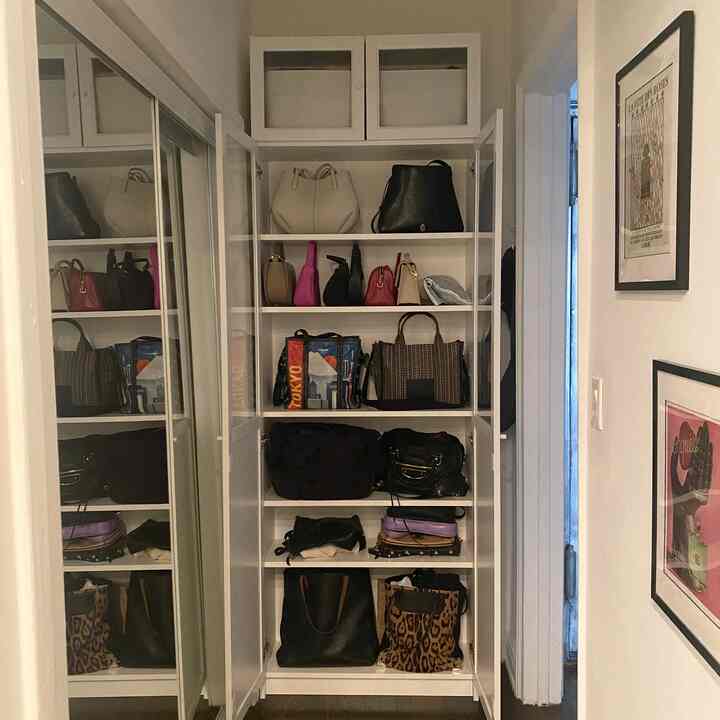 White-toned narrow and long corridor space featuring a tall organized shelving unit filled with various colored handbags and a large mirror on the side, creating a neat interior