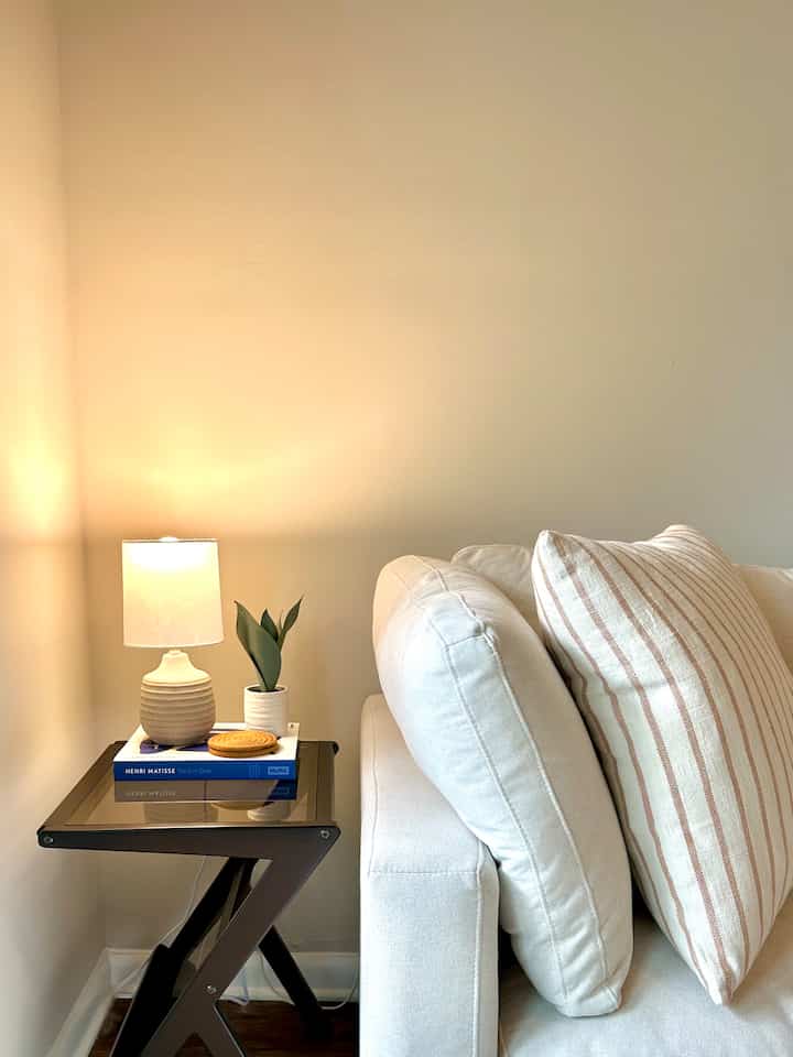 White and beige toned living room featuring a sectional sofa with cushions and a Z-shaped side table with a table lamp creating a cozy atmosphere
