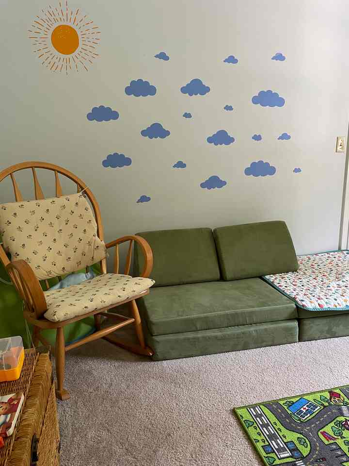 Natural color living room featuring an olive green sofa, wooden rocking chair, kids' play rug, and cheerful wall decals creating a cozy atmosphere