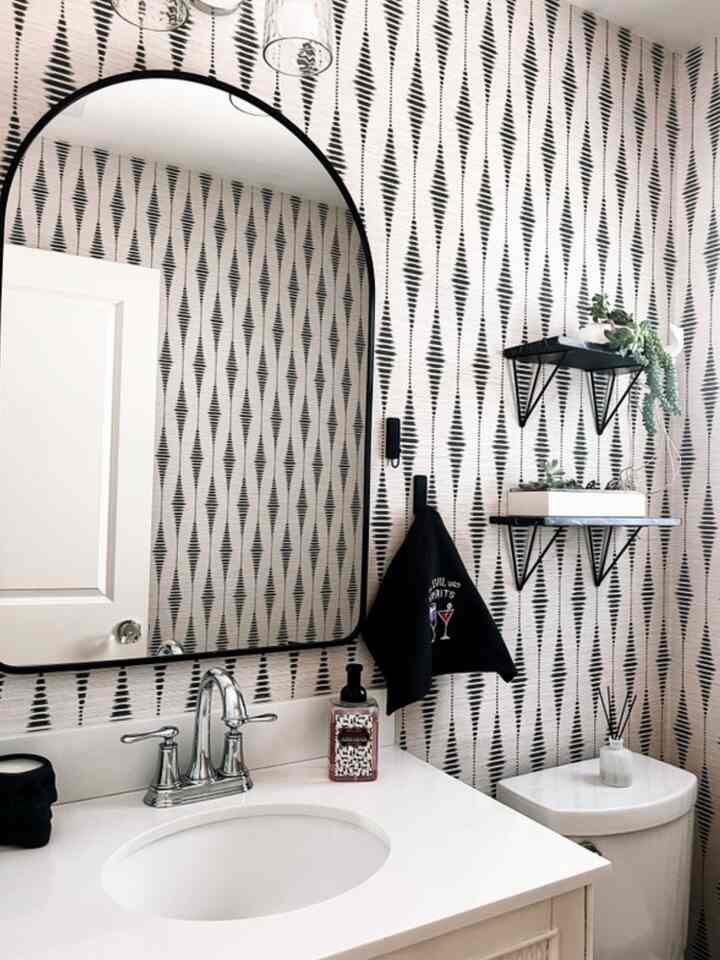 Black and white patterned wallpapered white bathroom featuring a vanity mirror, metal shelves, and plants creating a cohesive space