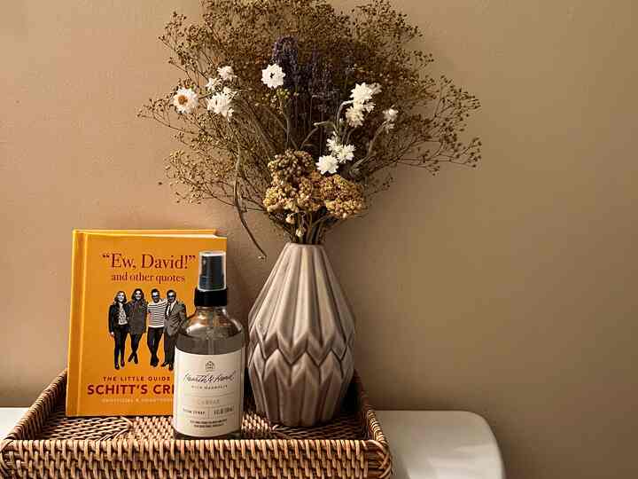 Beige wall with a natural bathroom essentials setup featuring dried flowers in a vase and diffuser on a rattan tray