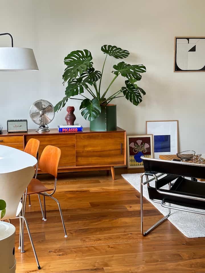 Dining room with white walls and wood-tone furniture, featuring a large green plant and modern chairs in a cozy setting