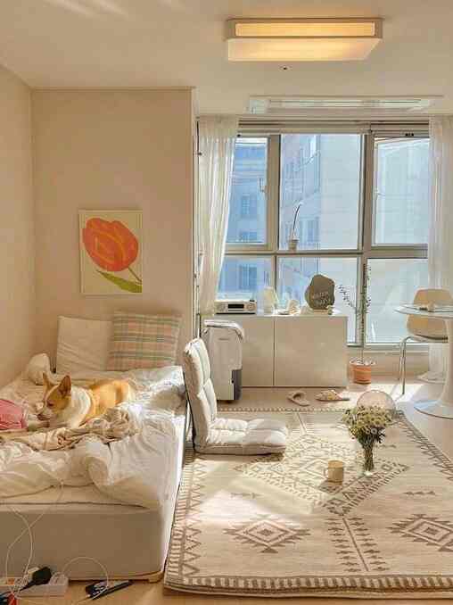 Cozy natural-toned bedroom featuring a bed, rug, and floor chair with bright light streaming through large windows