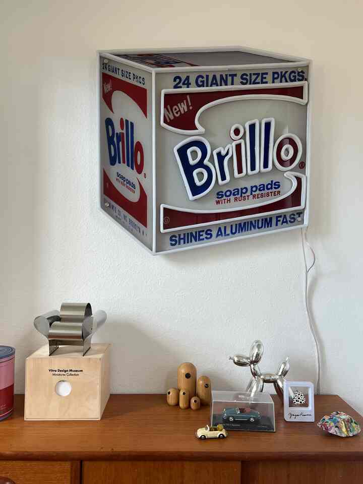White wall with Brillo wall decor and wooden shelf displaying art objects in a modern, artistic interior space