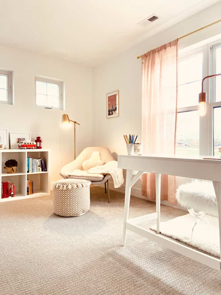 A soft beige and white toned living room featuring an armchair, woven pouf, white desk, and pastel pink curtains, creating a cozy atmosphere