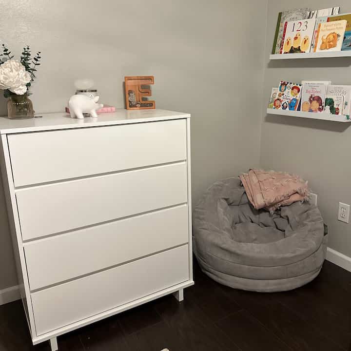 Cozy book nook in white and gray tones featuring a white dresser, floating shelves with books, and a gray bean bag chair