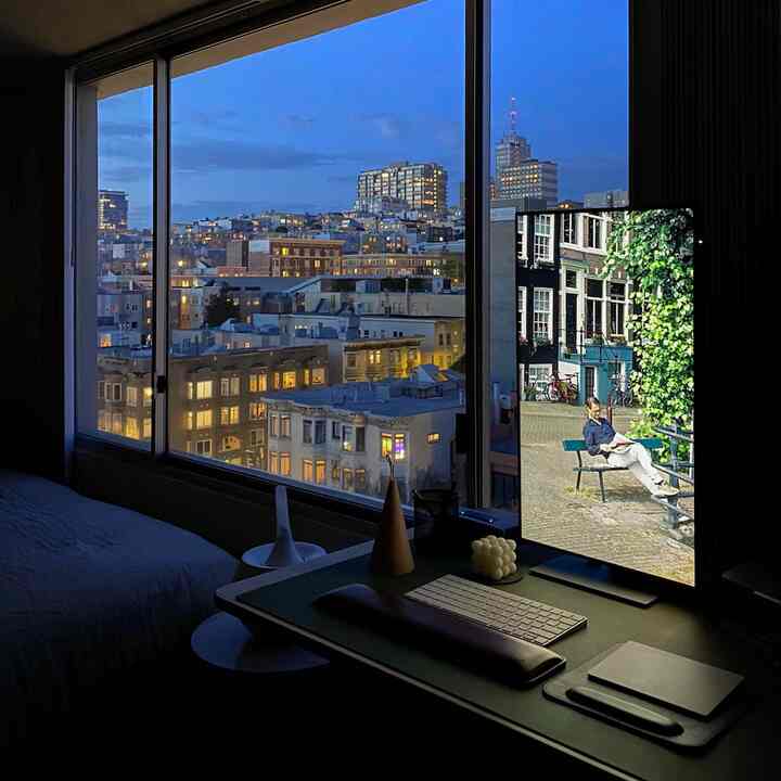 Dim blue-toned bedroom home office with desk setup facing large window showing city nightscape