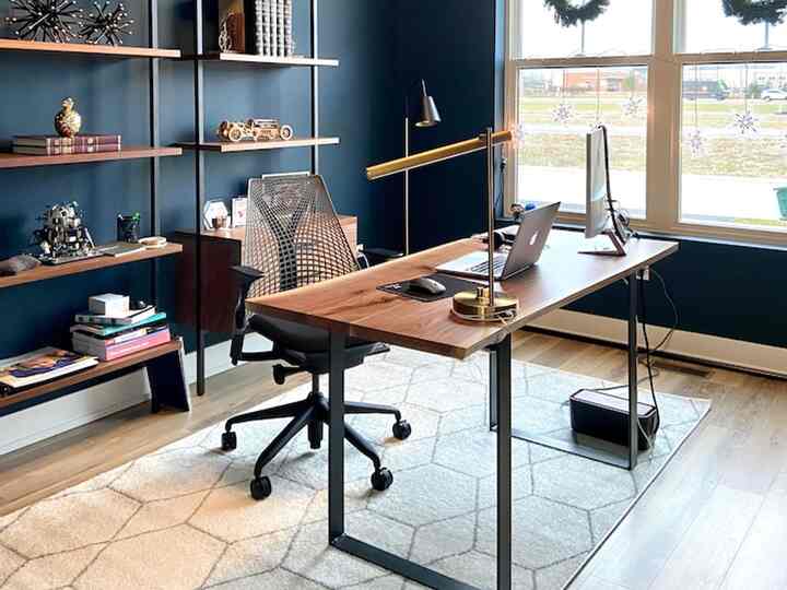 A modern home office with navy walls, wood-toned desk, black office chair, and walnut bookshelf featuring a bright atmosphere