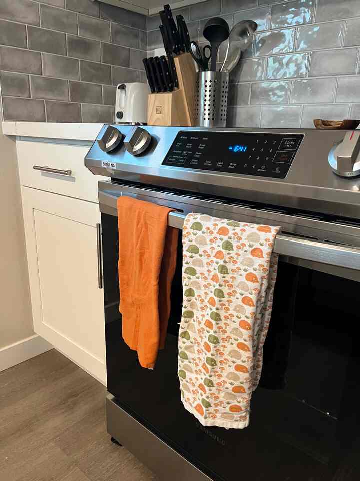 Modern kitchen featuring a stainless steel oven with orange and white towels hanging, white cabinets, and a tiled backsplash.