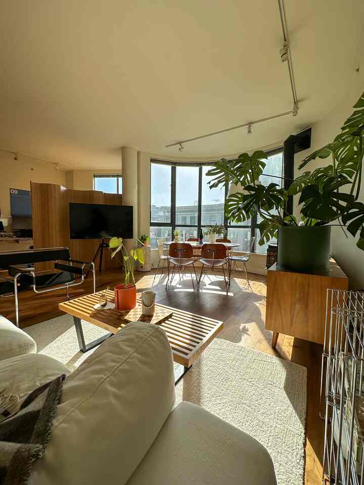 Bright living room with white walls and wood-toned furniture, featuring a dining table and plants creating a natural atmosphere