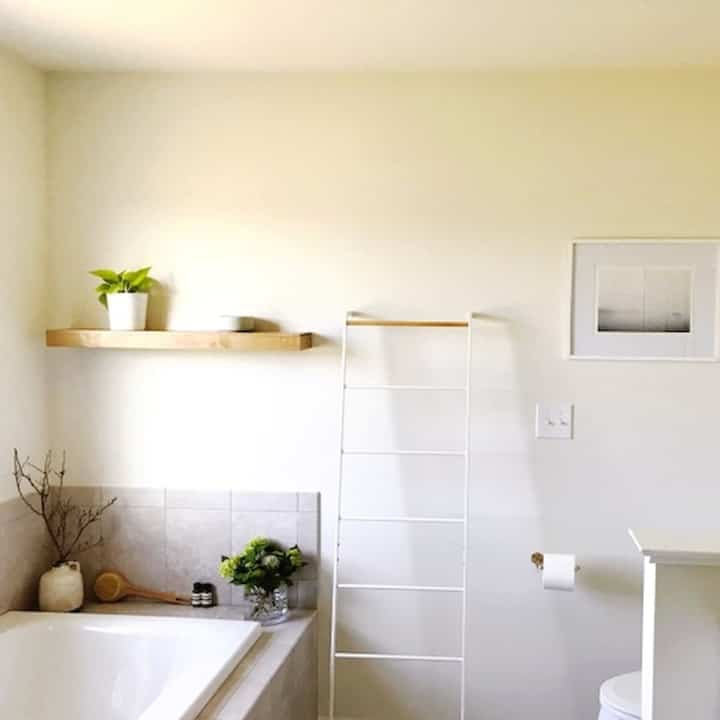 Bright white and natural color bathroom featuring wood floating shelf and metal towel rack in a clean minimalist design