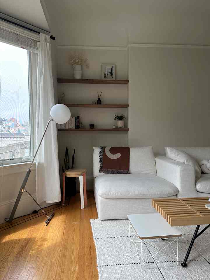 White and wood tone living room featuring white sectional sofa, wooden shelves, and floor lamp with a calm, peaceful interior vibe