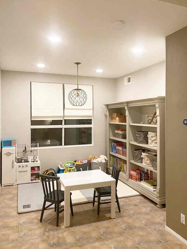 A white and natural-tone kids' room featuring a central kids table with chairs and a large bookshelf on the right, clean and organized space