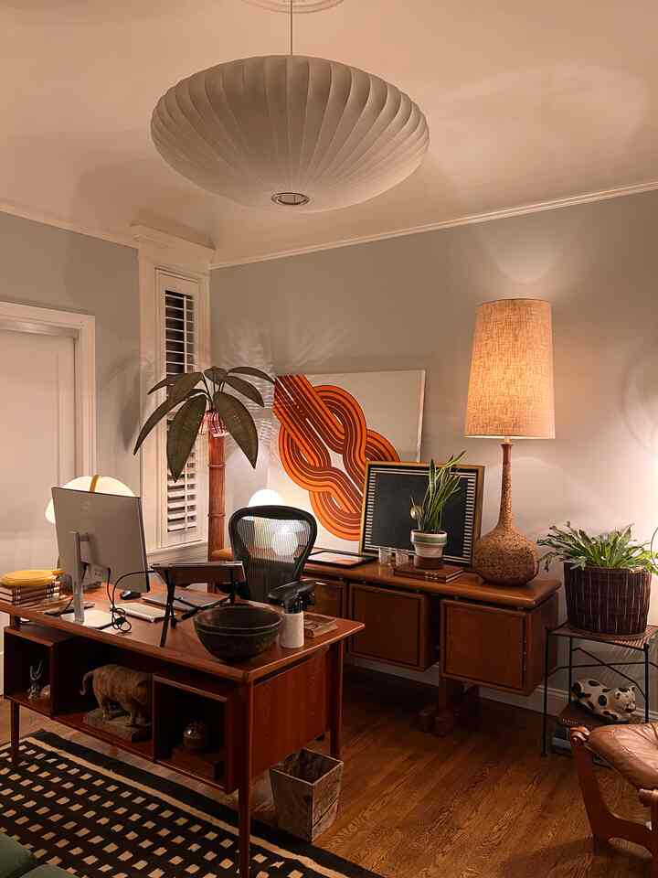 Warm wood-toned beige home office featuring mid-century modern furniture, floor lamp, plants, and artistic wall decor creating a cozy atmosphere