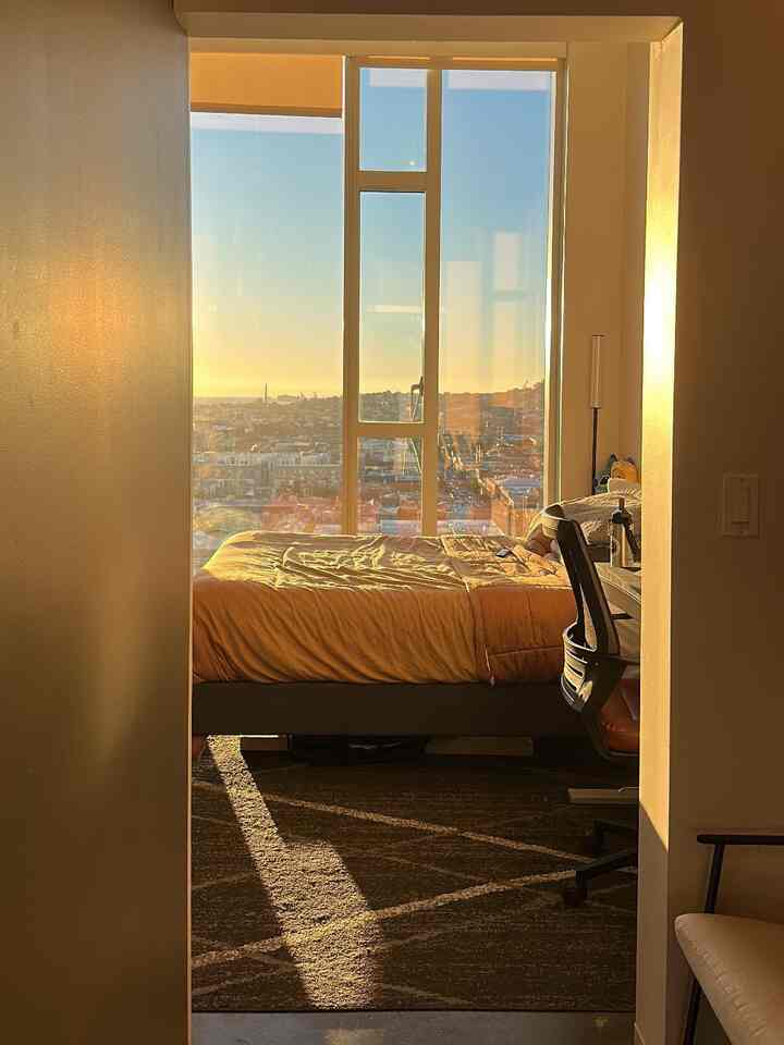 Golden-toned bedroom featuring large floor-to-ceiling window, bed frame, and a cozy home office setup with modern design