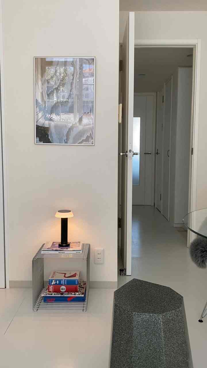Bright white-toned small indoor space featuring a silver metal stool with a table lamp placed on top and a poster hung on the wall in a modern interior