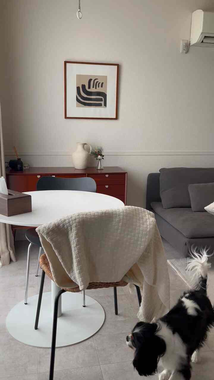 ベージュとグレーの色調のリビング、丸テーブルと木製キャビネットが配置されたシンプルで温かみのある空間、右下に犬がいるインテリア