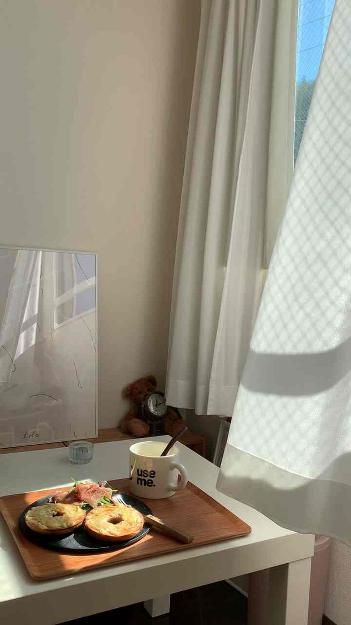 白いカーテンと木目調のトレイが置かれた6畳のワンルーム、サイドテーブル上の朝食が印象的な一人暮らしの雰囲気あるインテリア