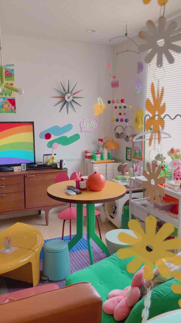 カラフルなリビング兼キッチン空間、中央に黄色の丸テーブルと緑のソファ、壁にレトロなサンバーストクロックが配置された印象的な空間