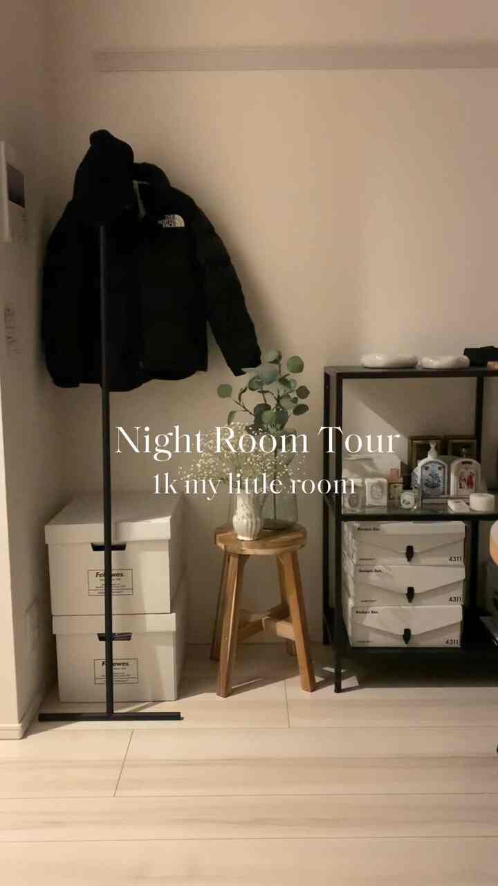 白い壁と床、黒い収納棚とコートラック、木製のスツールに植物が置かれたコンパクトな一人暮らしの空間