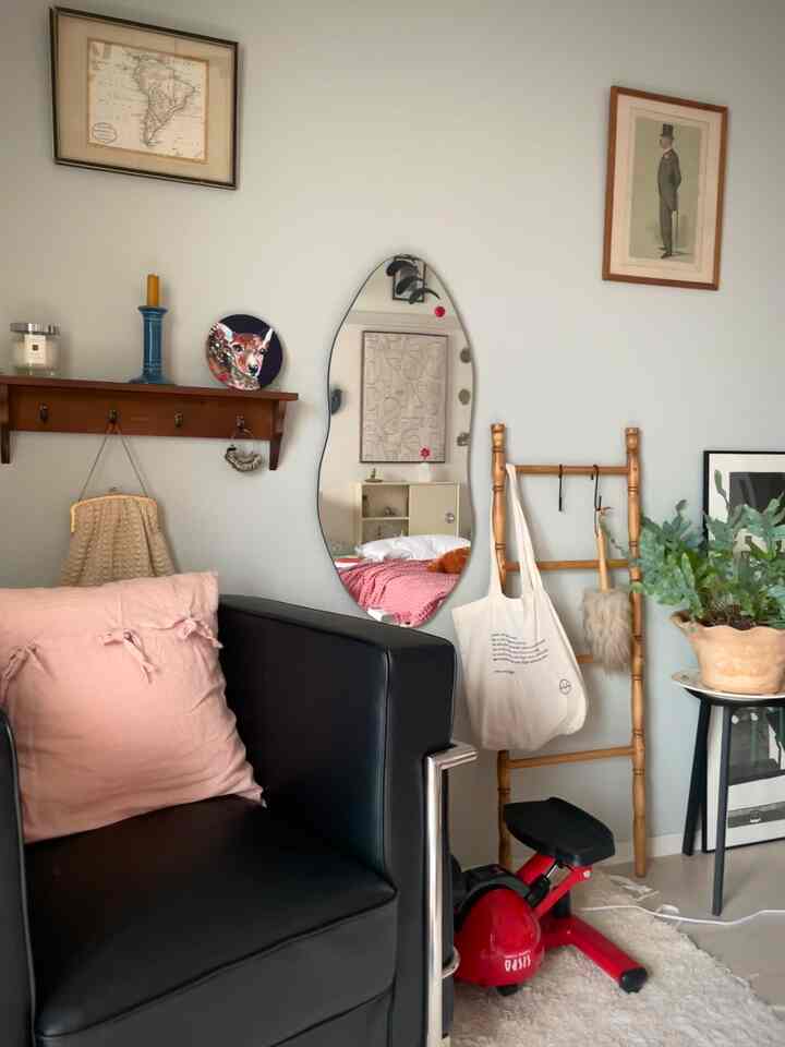 ナチュラルな色調の一人暮らしリビング、黒いレザーのアームチェアとピンクのクッション、特徴的な形の壁掛け鏡と植物が配置された居心地の良い空間