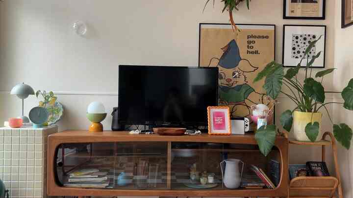 暖かく自然な色調のリビング、木製のテレビ台や複数の観葉植物、テーブルランプ、額縁が配置されたシンプルで印象的なインテリア