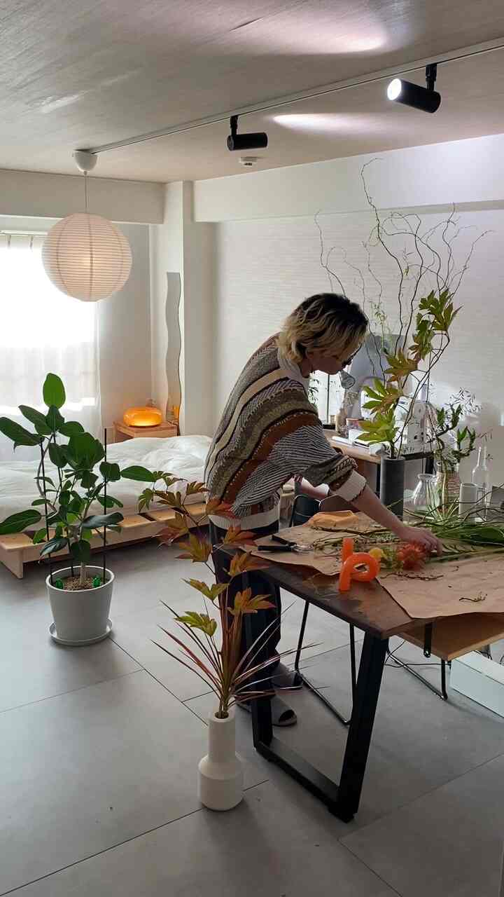 白とベージュのトーンのワンルーム、中央のテーブルで花を生ける人がいて、観葉植物とフロアランプが配置された自然な雰囲気のインテリア