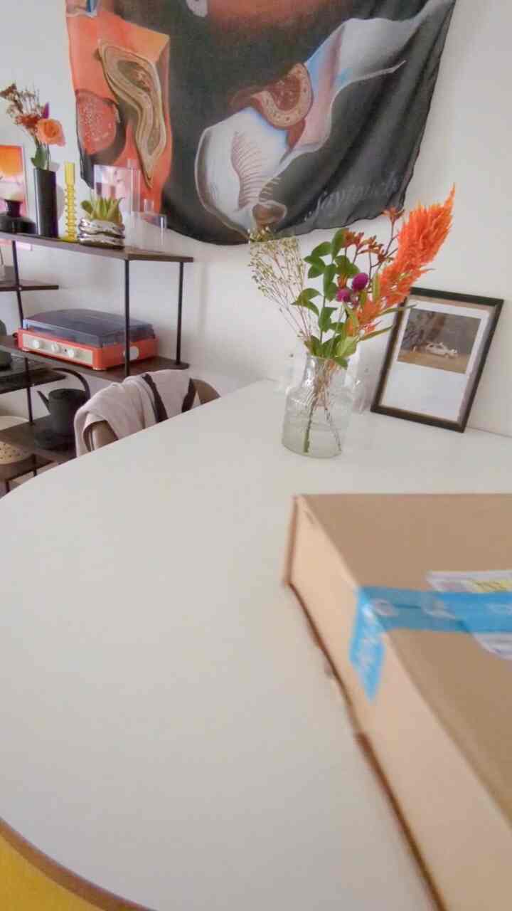 ナチュラルカラーのリビング、白いダイニングテーブルと花瓶、大きなファブリックのウォールデコが配置された雰囲気のあるインテリア