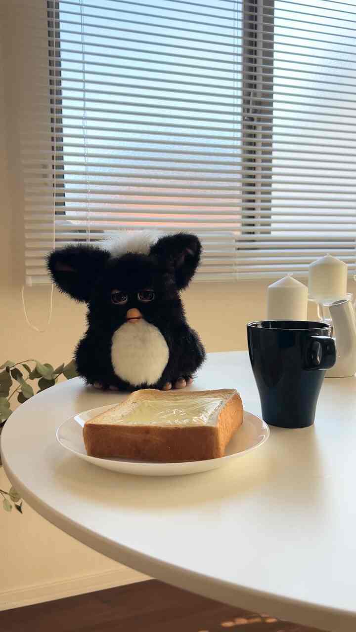 白を基調としたカフェ風のダイニングスペース、窓にはブラインドがあり、テーブルの上にトースト、マグカップ、ぬいぐるみが配置された温かみのあるインテリア