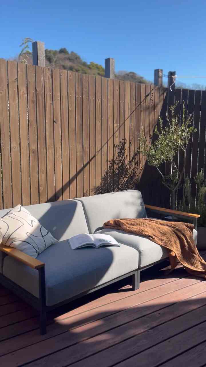 木目調と自然光が調和するウッドデッキのアウトドアリビングスペース、グレーのソファにクッションとオレンジのスローが配され、居心地の良い読書空間