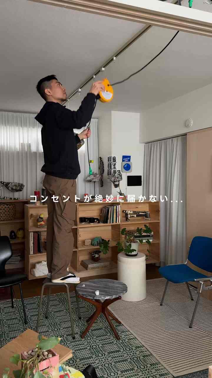 ナチュラルな色調の狭い一人暮らしのリビング、木製の本棚とラグ、そしてスツールに立つ人がいる印象的な空間