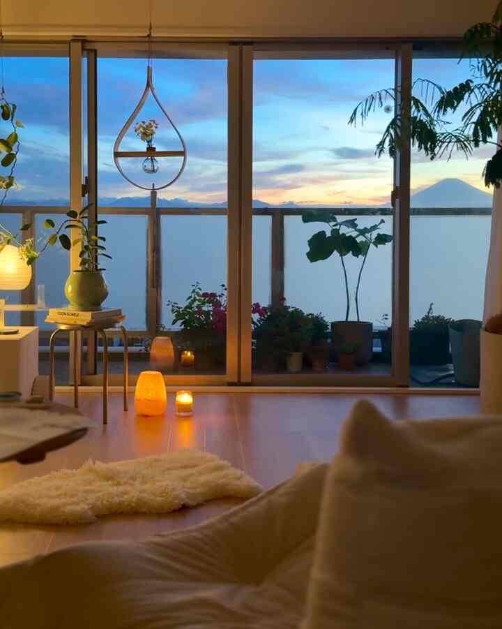Natural wood-tone living room with various plants and balcony visible through large window, warm candles creating cozy atmosphere
