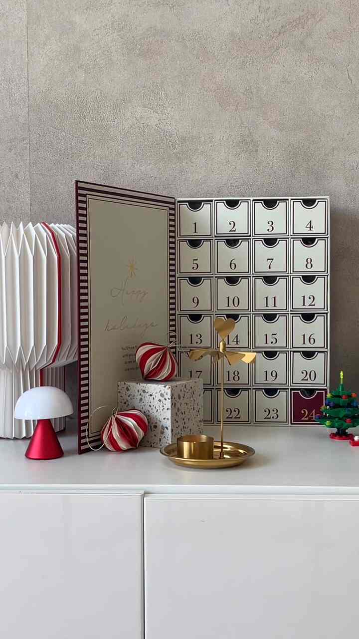白と赤のトーンでまとめられた棚の上に、クリスマスのアドベントカレンダーとテーブルランプ、ゴールドの飾りが配置された印象的なインテリア