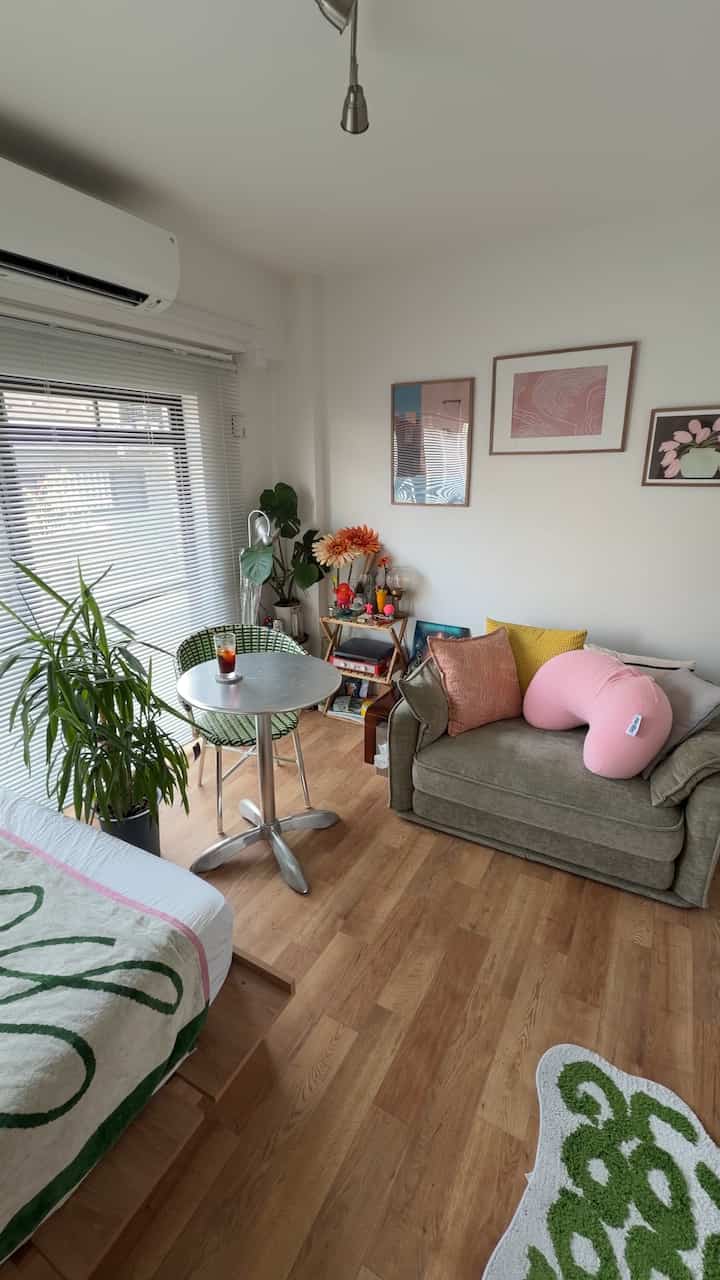 White-walled studio apartment living room with brown wood flooring, a green sofa, small round coffee table, plants, and framed artwork, creating a simple modern vibe