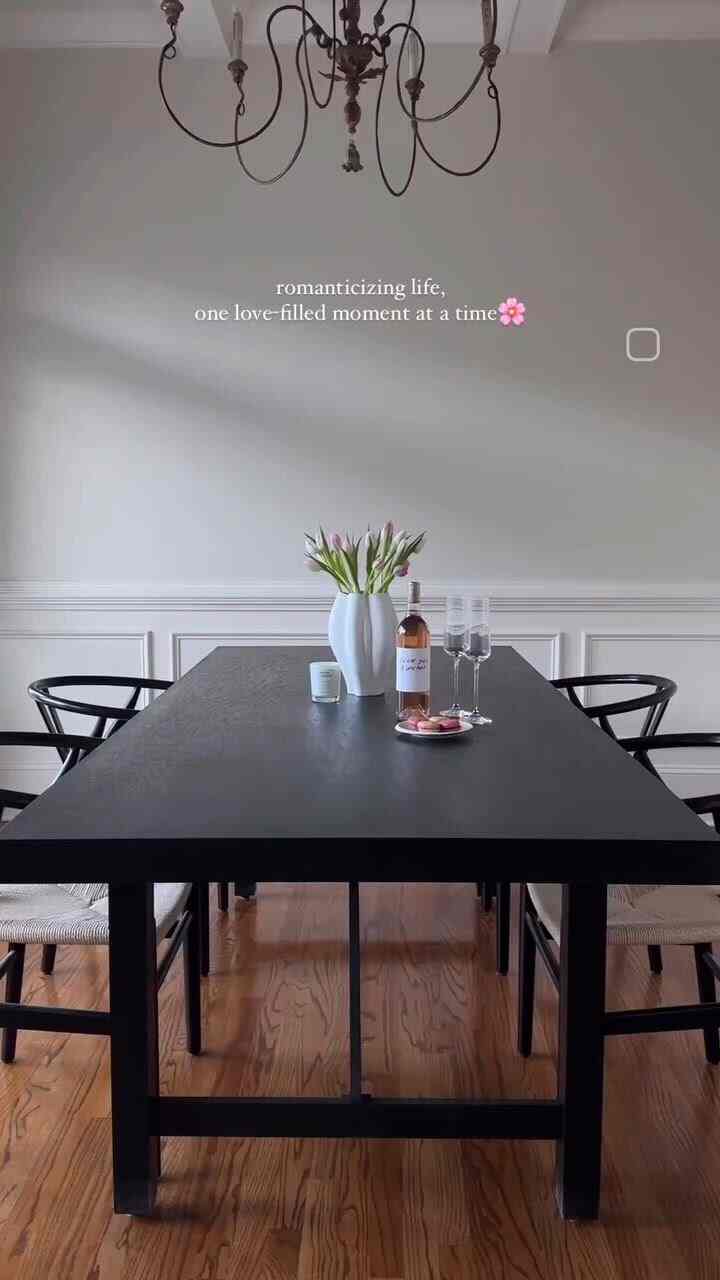 Natural-toned simple dining room featuring black table and chairs centered, with white vase of flowers, candle, and wine bottle on table