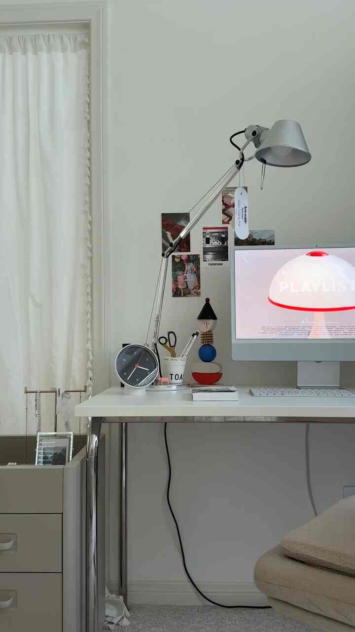 White and gray toned home office featuring a white desk with a silver table lamp, clock, and computer monitor in a clean setting