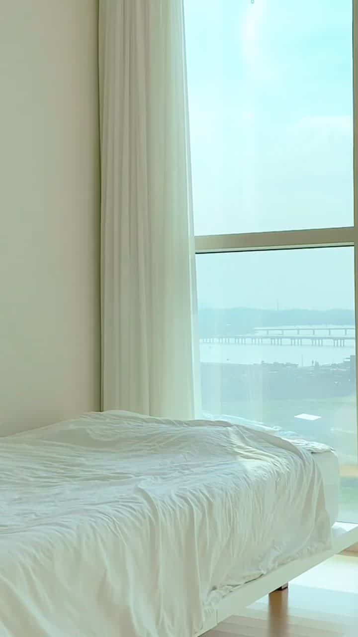 A minimal studio bedroom in white and natural tones featuring a large window and white curtains