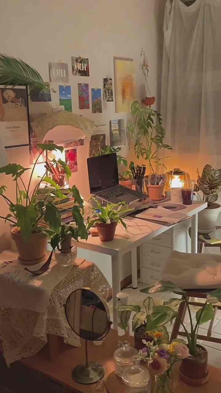 暖かい照明と白を基調としたワンルームのホームオフィス、多彩な植物と額入りポスターが配置された雰囲気のあるインテリア