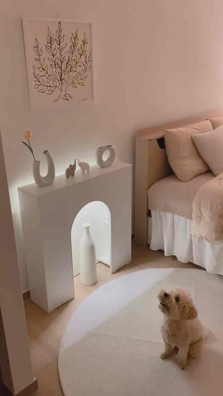 Cozy bedroom with white walls and light brown bed, featuring a fireplace console and a small dog sitting on a round rug