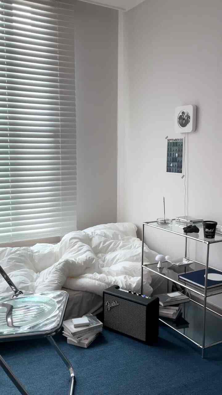 A modern studio bedroom featuring white walls, blue carpet, steel shelving, and a cozy duvet