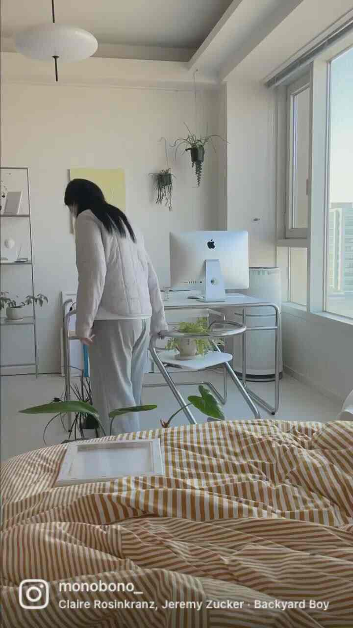 白を基調としたワンルーム、窓際に置かれたデスクとチェア、ストライプの布団カバーと植物が調和するモダンなインテリア