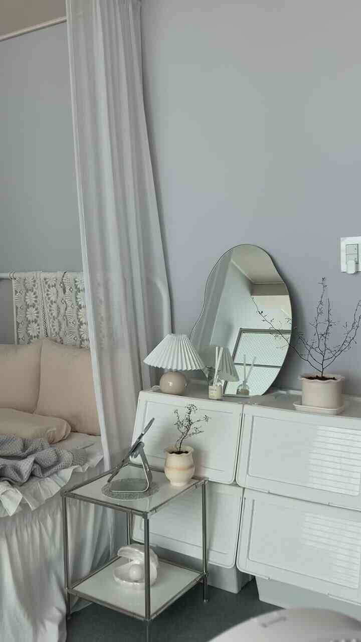 Small bedroom with gray-toned walls and white furniture, featuring a nightstand with lamp and plants in a minimal setting