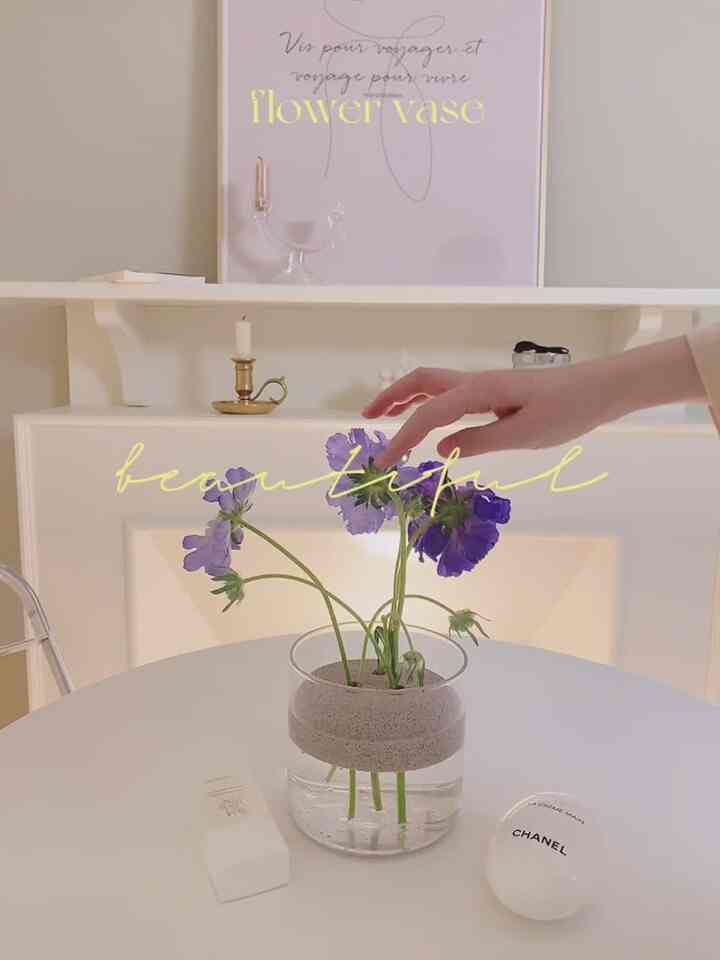 A white and natural color toned bedroom featuring a round table with a glass flower vase and purple flowers, creating a simple and cozy atmosphere