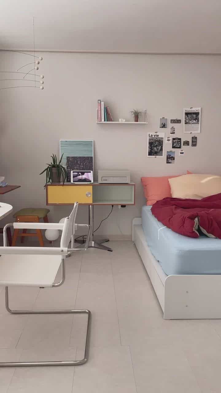White-toned studio bedroom featuring a modern desk and shelf, with comfortable bed and cushions in a cozy setup