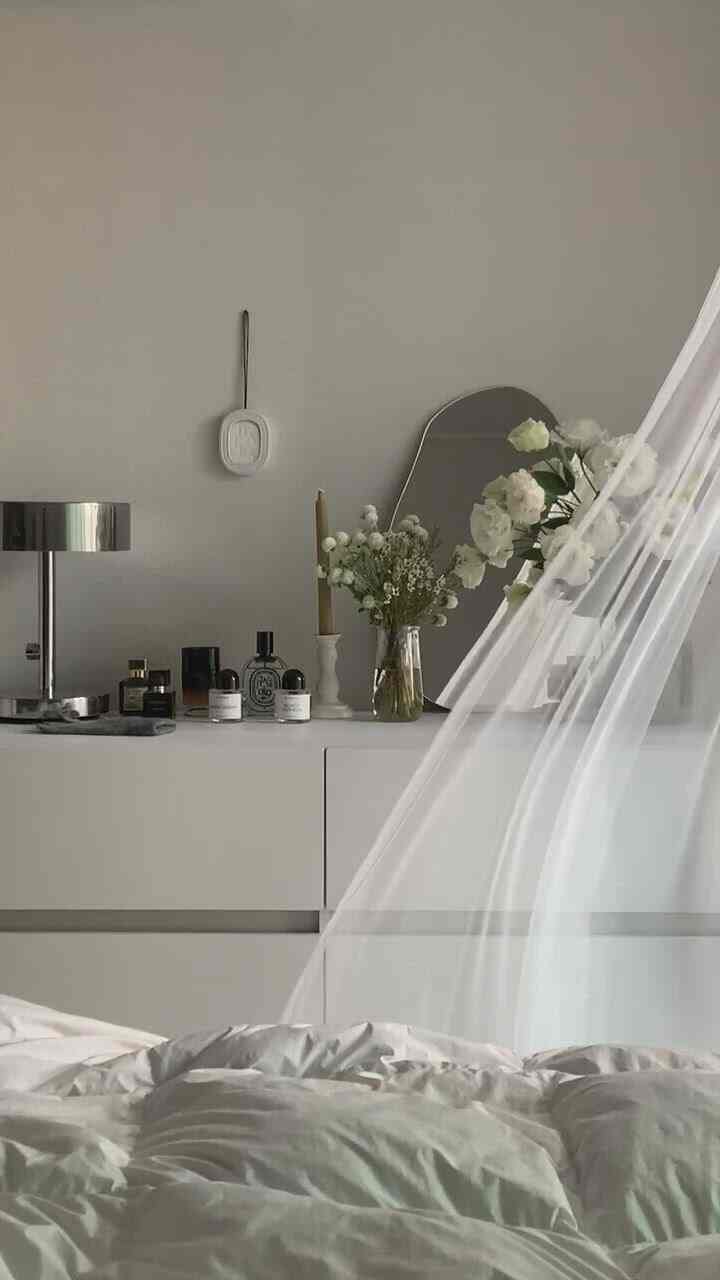 White-toned bedroom featuring a white bed, asymmetrical mirror, vase with flowers, and candle holder creating a calm natural atmosphere